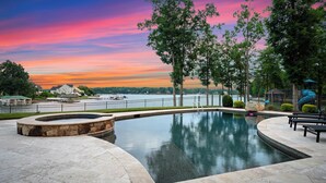 A swimming pool with a stone border, surrounded by a paved deck, and overlooking a body of water with a sunset sky. - Private Dock, Pool + HotTub, Game Room, Firepit | | Paradise Pointe by AvantStay (Sherrills Ford)