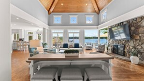 A spacious living area with a wooden ceiling, large windows, a stone fireplace, and a TV mounted on the wall. - Private Dock, Pool + HotTub, Game Room, Firepit | | Paradise Pointe by AvantStay (Sherrills Ford)