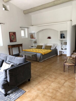 Quarto com cama, sofá, poltrona, mesinha e prateleira com livros e objetos decorativos.
