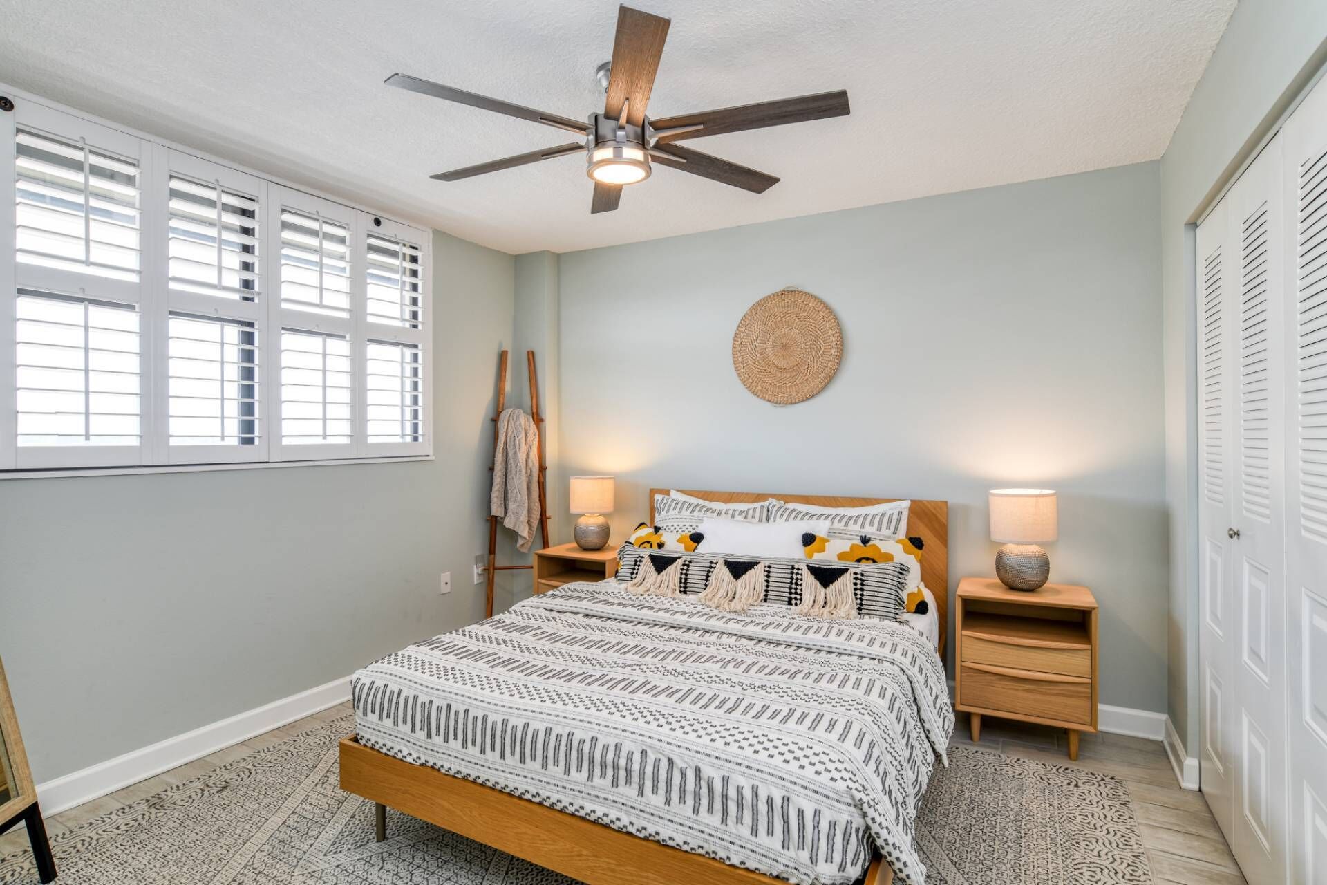 A bedroom with a bed, two bedside tables, a ceiling fan, a window with blinds, and a door.