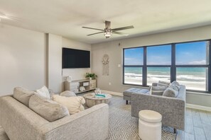 A living room with a large window overlooking the beach, a ceiling fan, and a flat-screen TV mounted on the wall.