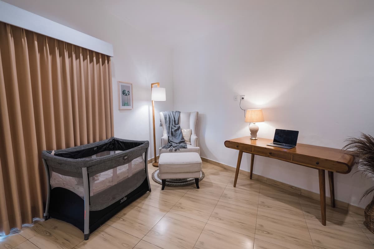 A room with a convertible crib, a white chair, a wooden table with a laptop, and a lamp.