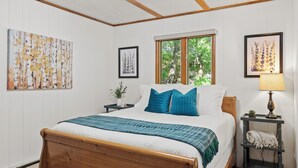Un dormitorio con cama de madera, paredes blancas y ventana con vistas a árboles.