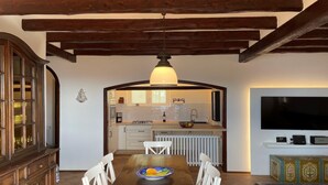 Uma sala de jantar com mesa e cadeiras de madeira, uma cozinha ao fundo e uma televisão de tela plana na parede.