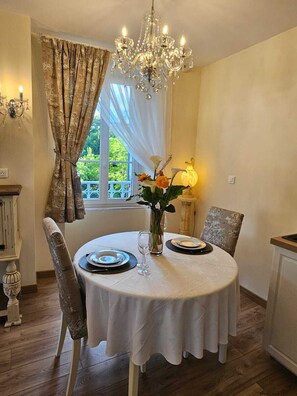 Un coin repas avec une table ronde dressée pour deux personnes, un lustre et une vue sur la verdure environnante.