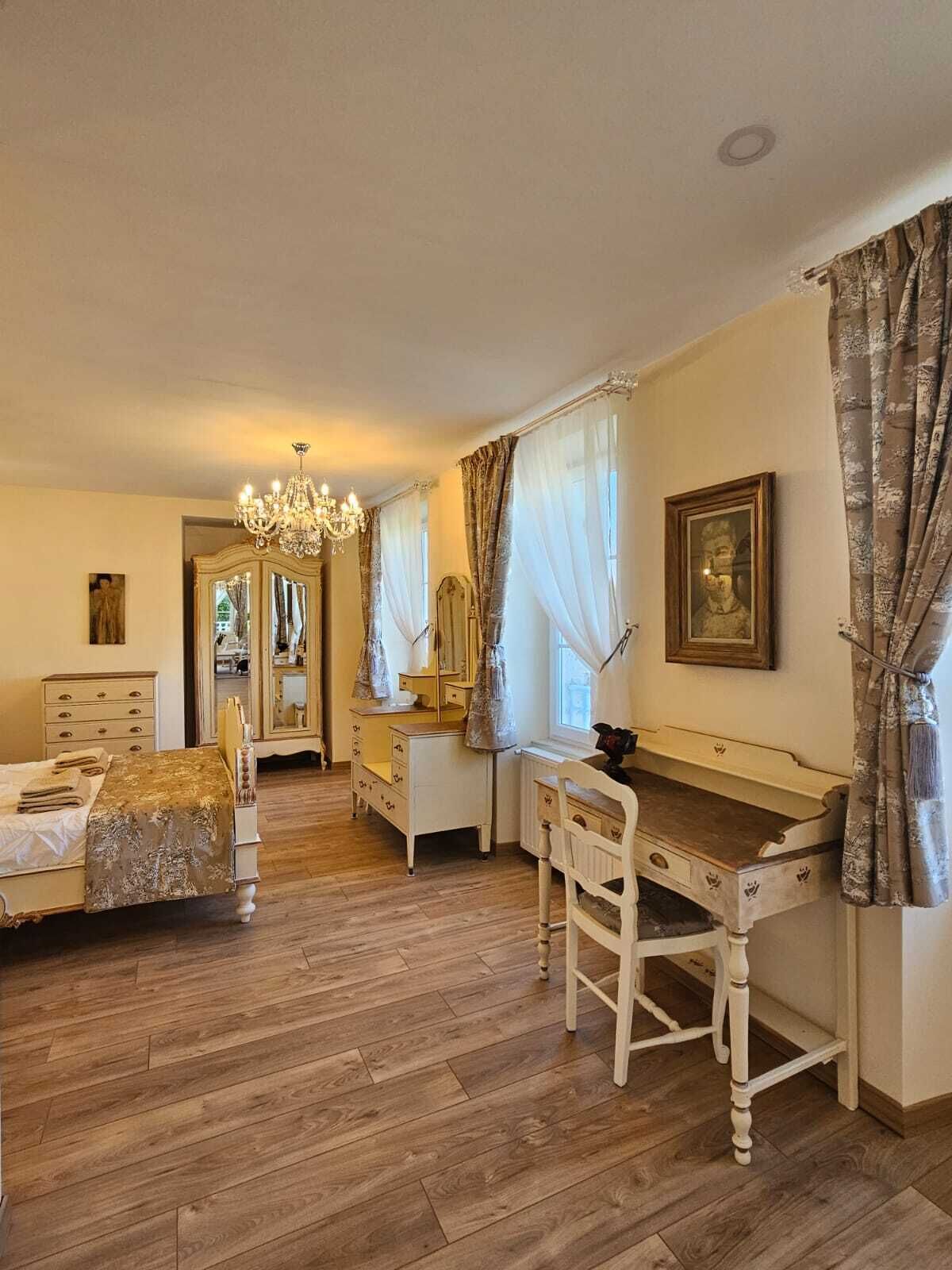 Une chambre avec un plancher en bois, un lit recouvert d’une couverture dorée, une commode, une coiffeuse avec un miroir et un bureau avec une chaise.
