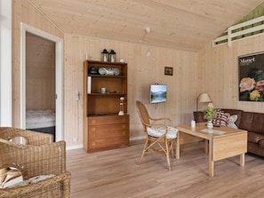 Una habitación con suelo de madera, una cama, una estantería de madera, un sillón de mimbre, una mesa, un sofá de cuero marrón y un televisor montado en la pared.