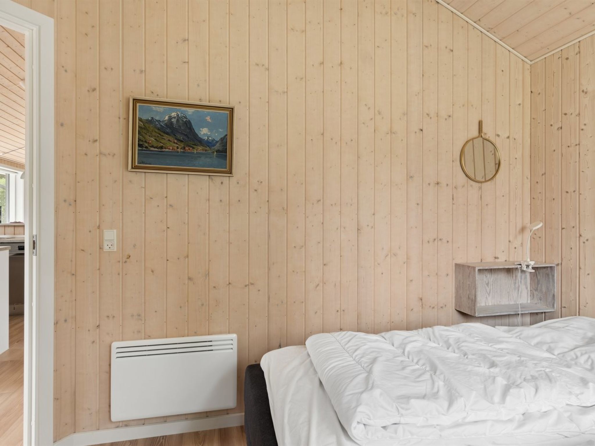 A room with wooden panel walls, a bed with white bedding, a mounted picture, a mirror, and a small wooden shelf.