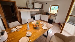 A dining area with a wooden table set for four, a kitchen in the background, and a window with blinds.