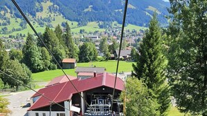 A mountain lodge with a red roof, a ski lift, and a view of a green valley and mountains in the background.
