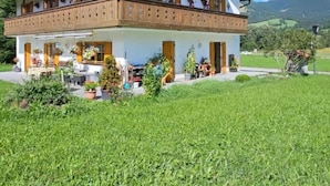 A two-story chalet with a balcony, surrounded by a well-maintained lawn and a mountainous backdrop.