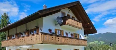 A two-story chalet with a balcony, surrounded by a well-maintained lawn and a mountainous backdrop.