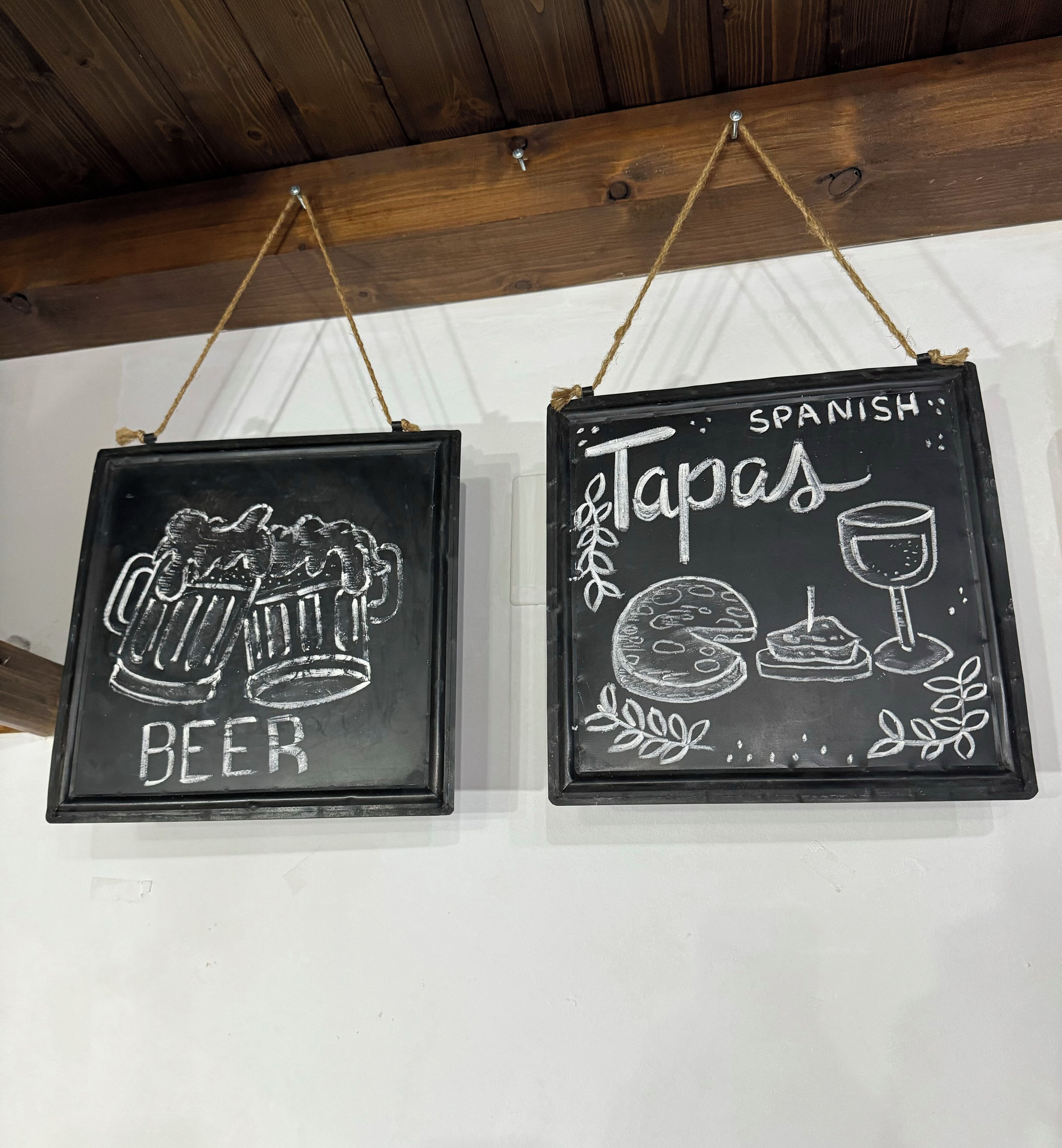 Two black chalkboard signs with white drawings and text, hanging from a wooden beam.