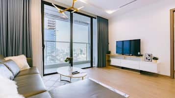 A modern living room with a sofa, a coffee table, a TV on a white cabinet, and a large window with a city view.