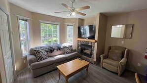 A living room with a sofa, a coffee table, a fireplace, a television, and a ceiling fan. - 3 Story Riverside  Townhome (Fernie)