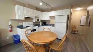A compact kitchen with white cabinets, a stove, microwave, and refrigerator. There is a wooden dining table with four chairs. - 3 Story Riverside  Townhome (Fernie)