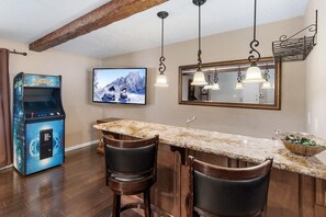 A kitchen with a marble countertop, two bar stools, a built-in bar, a television mounted on the wall, and a retro arcade game. - MJ's Hideaway By Destination Big Bear: Walking Distance to Snow Summit!Game Room! Hot Tub! Pet Friendly! Fenced Yard! (Big Bear Lake)