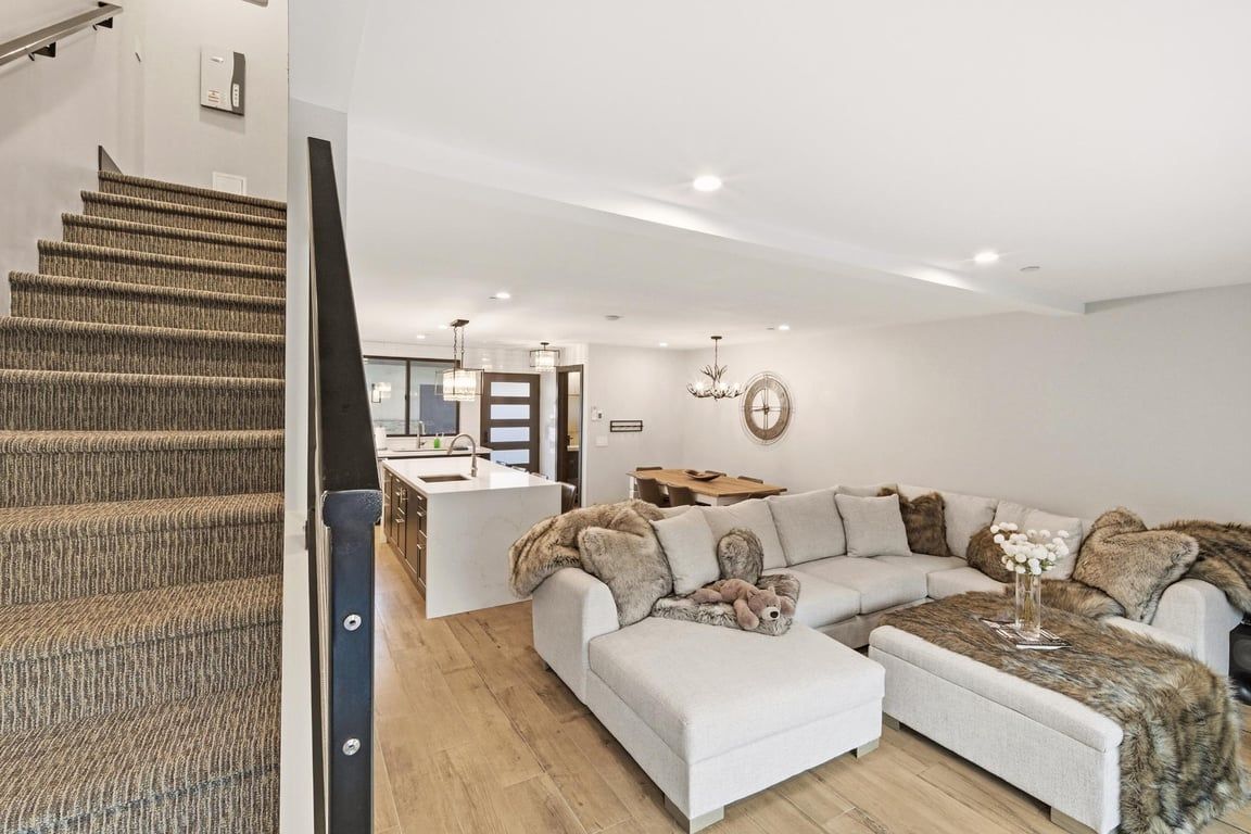 A modern interior with a staircase, a living area with a sofa and ottoman, a kitchen area, and a dining table.