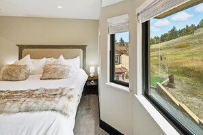 A bedroom with a large bed, a nightstand, a lamp, and a window with a view of a grassy landscape. - Clip In and Ride Straight Onto Chair 9 at Bear Mountain! Private HOT TUB! Air Conditioning! Pickle Ball Court! EV Charger! (Big Bear Lake)