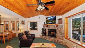 A cozy living room with a stone fireplace, wooden ceiling, and a large window overlooking a deck.