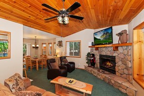 A cozy living room with a stone fireplace, a ceiling fan, and a television. - Freddy's Lake House: Walking Distance to Lake and Meadow Park! Fenced Yard! Spa! (Big Bear Lake)