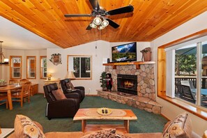 A cozy living room with a stone fireplace, a television, and a dining area with wooden furniture. - Freddy's Lake House: Walking Distance to Lake and Meadow Park! Fenced Yard! Spa! (Big Bear Lake)