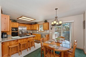 A kitchen with wooden cabinets, a stainless steel refrigerator, a dining table with chairs, and a hanging light fixture.