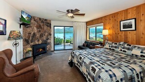A bedroom with a stone fireplace, a bed with a checkered blanket, a ceiling fan, and a view of a lake through a large window. - Lakefront Haven:Lakefront Condo! Located in the Village! Community Spa & Pool! Private Dock! (Big Bear Lake)