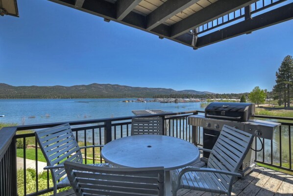 A deck with a round table and metal chairs, overlooking a lake with mountains in the background.