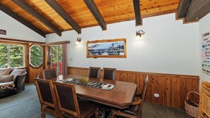 A dining area with a wooden table and chairs, a painting on the wall, and a view of the outdoors through large windows.