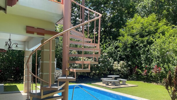 Ein Poolbereich mit einer Treppe, die zu einer überdachten Terrasse führt, umgeben von üppiger Vegetation.