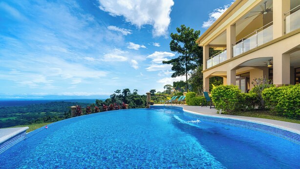 Una grande piscina azzurra e limpida con bordo curvo, immersa nel verde e affiancata da un edificio con balconi.