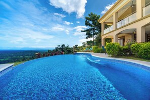 Une grande piscine à l’eau bleue et limpide, à bords incurvés, entourée d’une végétation luxuriante et bordée d’un bâtiment doté de balcons.