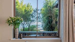 A room with a large window, a bed, a bedside table with a vase of flowers, and a view of trees and a body of water.