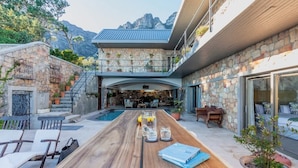 A modern outdoor dining area with a pool, stone walls, and a mountain view.