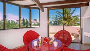 Une pièce avec une table en verre, des chaises en osier rouge et une vue sur des palmiers et des immeubles.