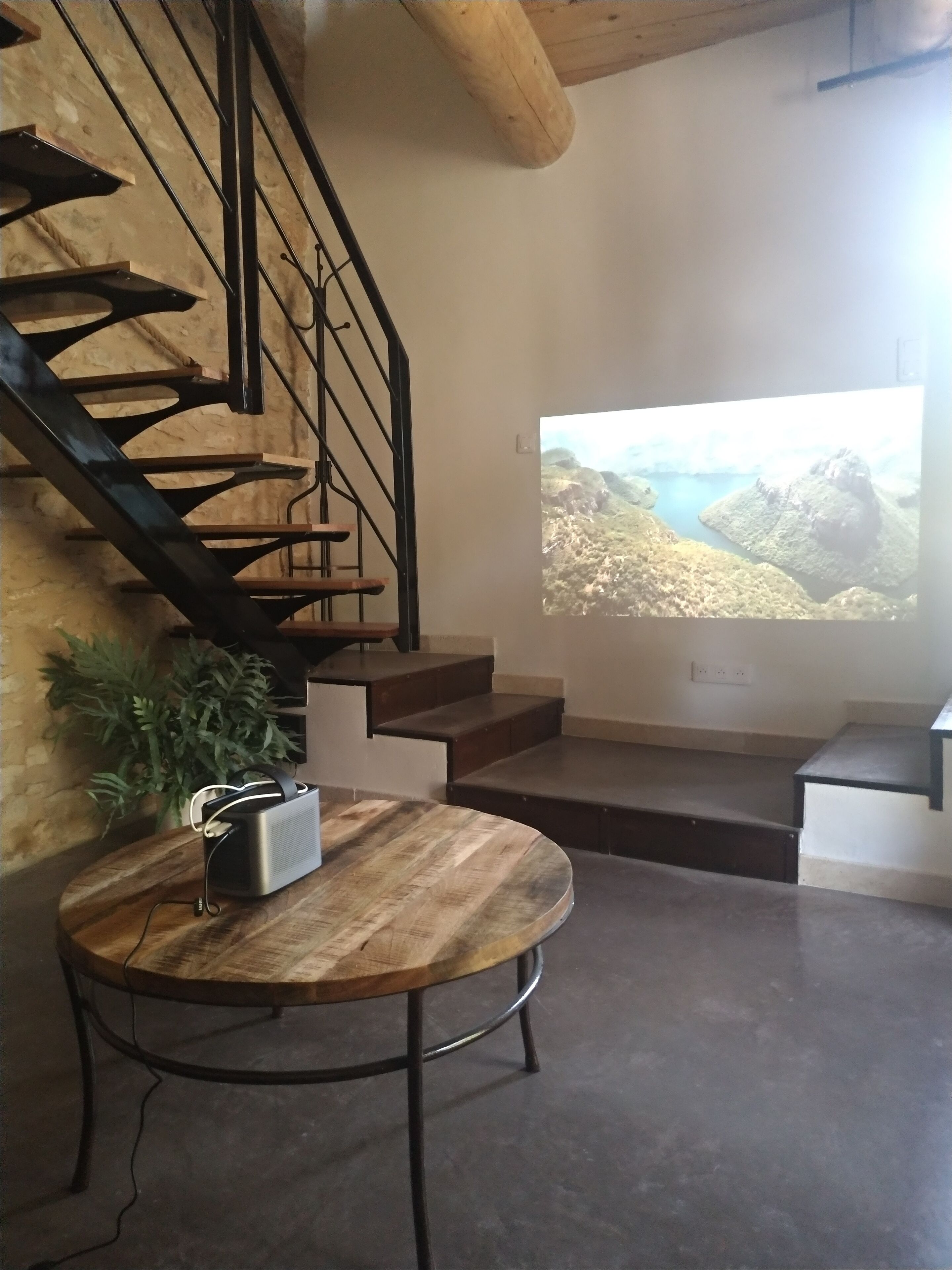 A modern living room with a wooden staircase, a large TV screen displaying a scenic view, and a round wooden table with a speaker.
