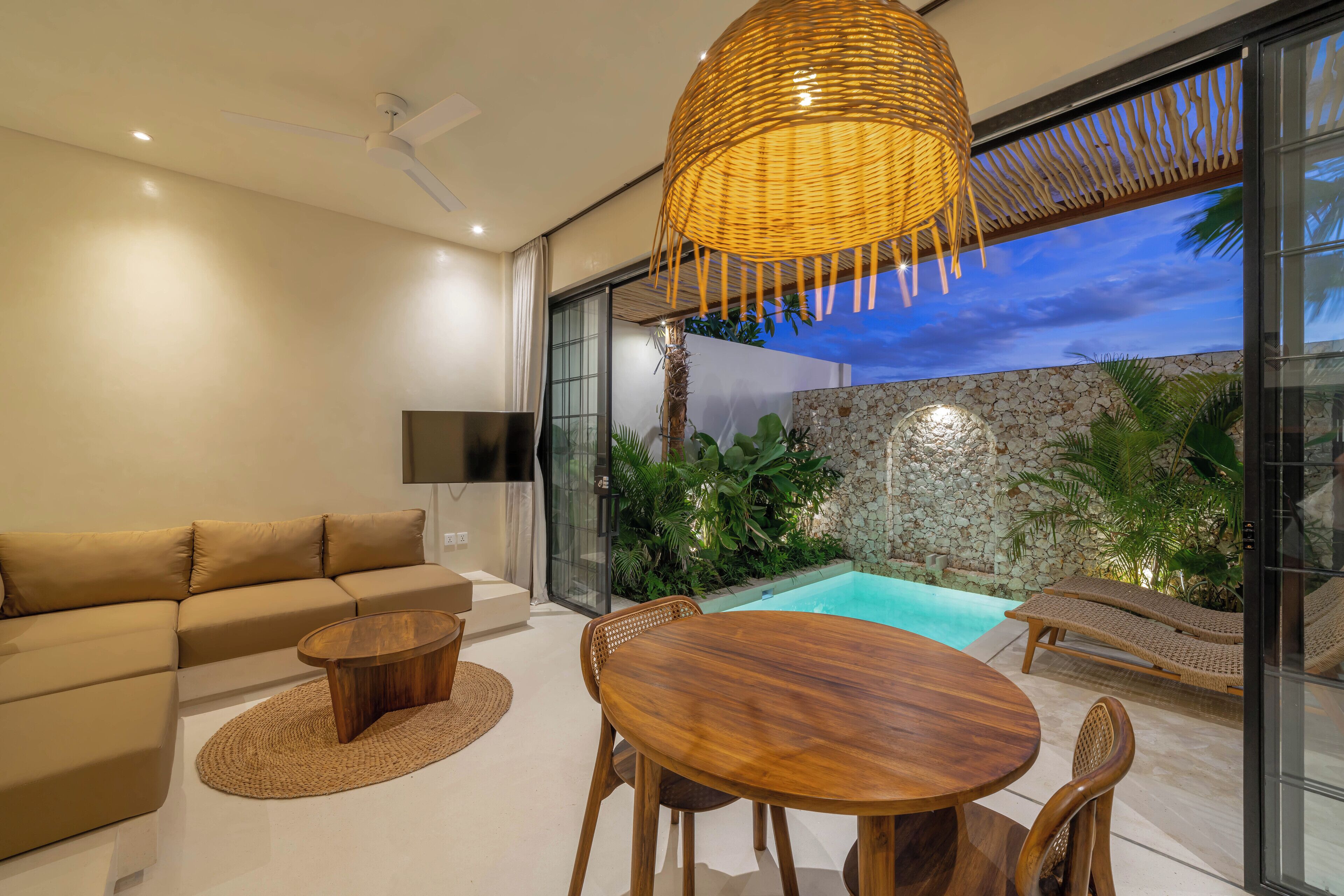 A modern living room with a sofa, coffee table, and dining table. There is a large window with a view of a swimming pool and greenery.