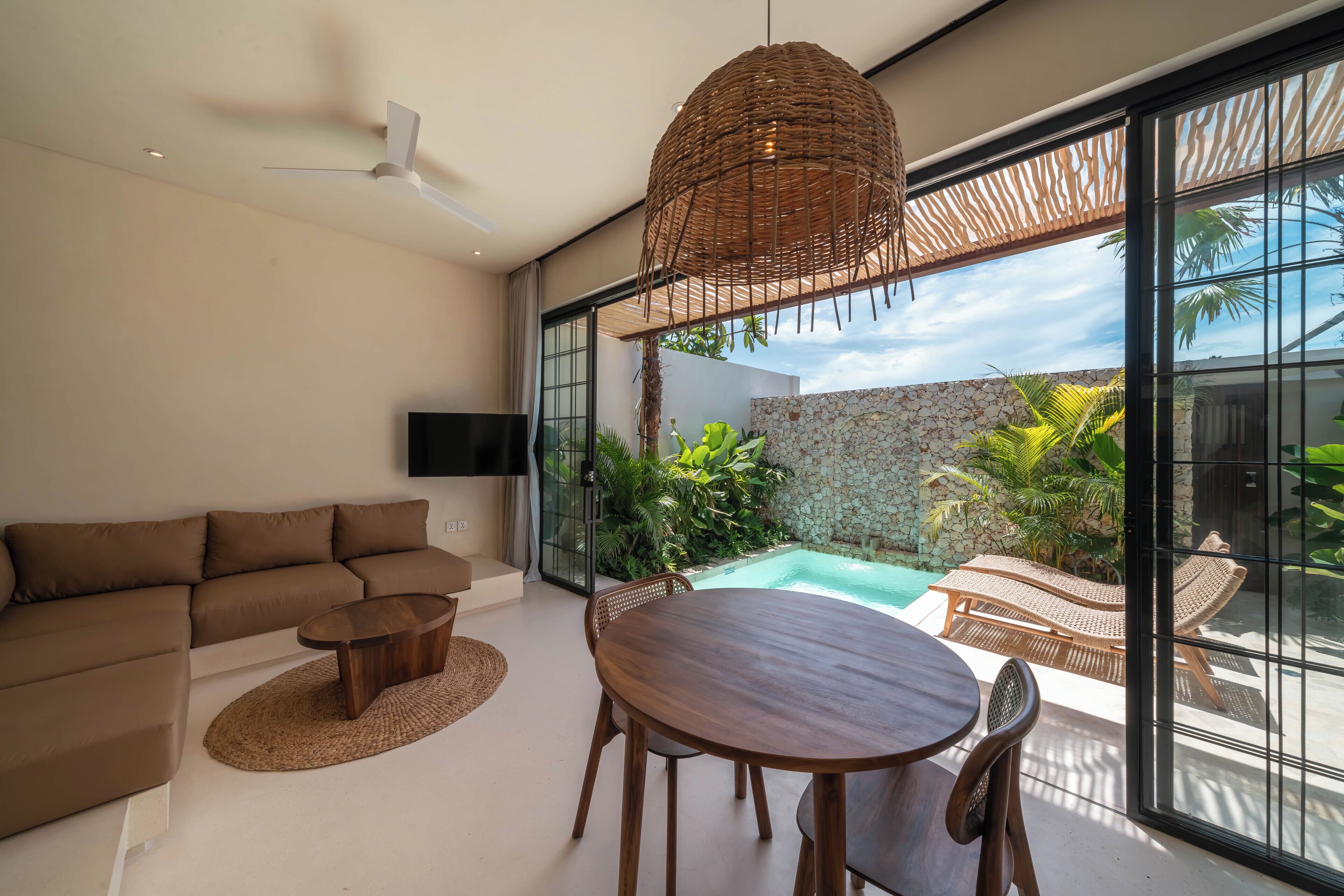 A modern living room with a sofa, a wooden table, and a TV. There is a large window with a view of a swimming pool and greenery.