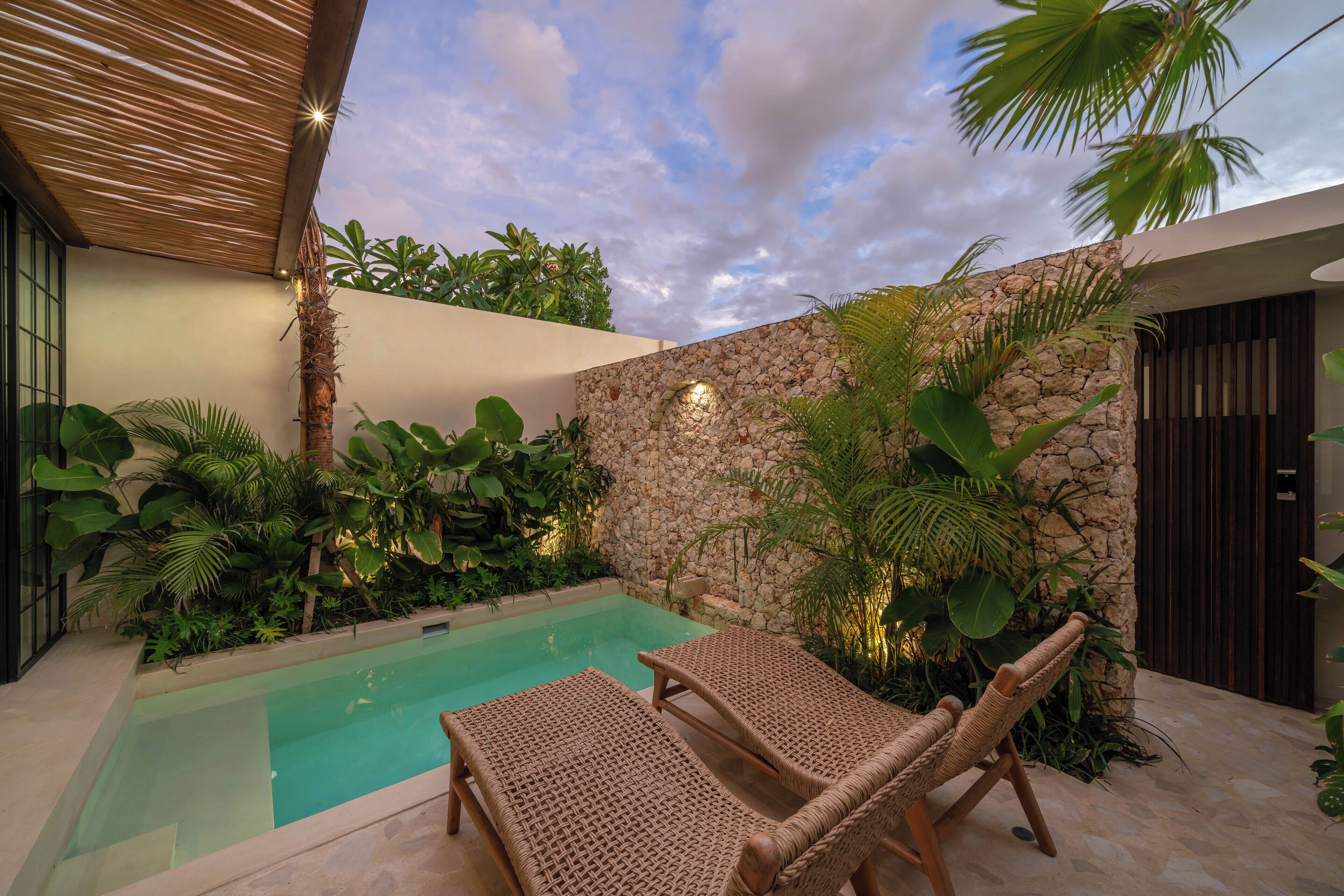 A pool area with two wicker lounge chairs, a stone wall, and lush tropical plants.