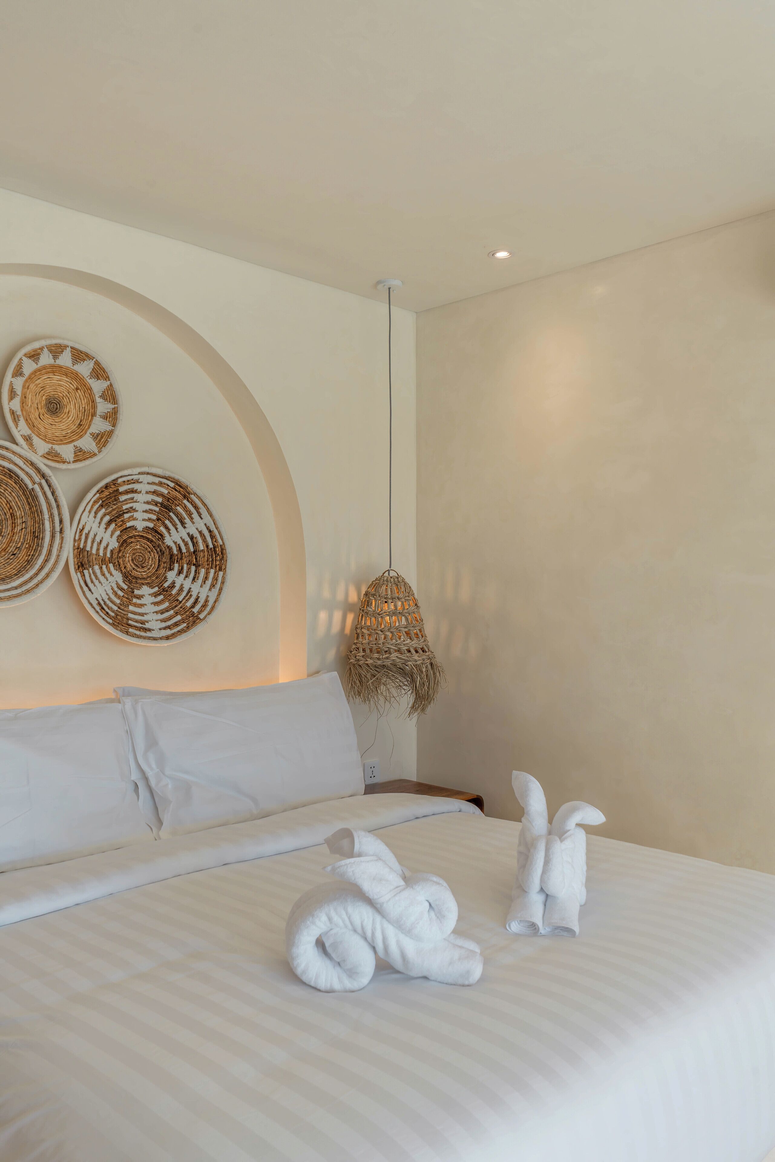 Habitación de hotel con una cama grande, ropa de cama blanca, tres piezas de arte de pared tejidas y una lámpara colgante de fibra.