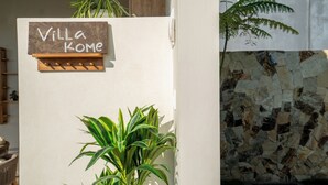 Bordje met de tekst 'Villa Kome' op een muur, een bloempot en een stenen muur met vuurkorf.