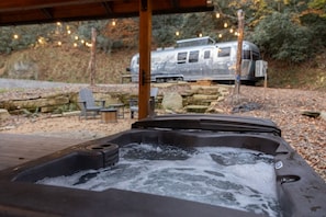 A hot tub with a wooden deck and stone walls, a chair, and a camper in the background.
