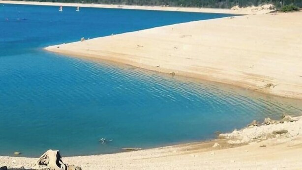 Une plage avec une eau d’un bleu limpide, un rivage sablonneux et une colline boisée en arrière-plan.