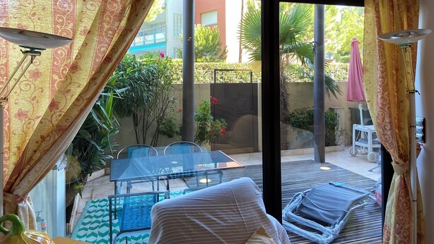 A room with a sliding glass door leading to an outdoor patio area. The patio has a table and chairs, a lounge chair, and a small table with a lamp. - Family duplex overlooking the beach (Antibes)