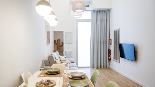 Un comedor con una mesa preparada para dos personas, un sofá al fondo y un televisor montado en la pared.
