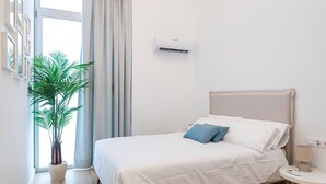 Un dormitorio ordenado con cama, una mesita de noche con lámpara, una planta en maceta y un equipo de aire acondicionado en la pared.