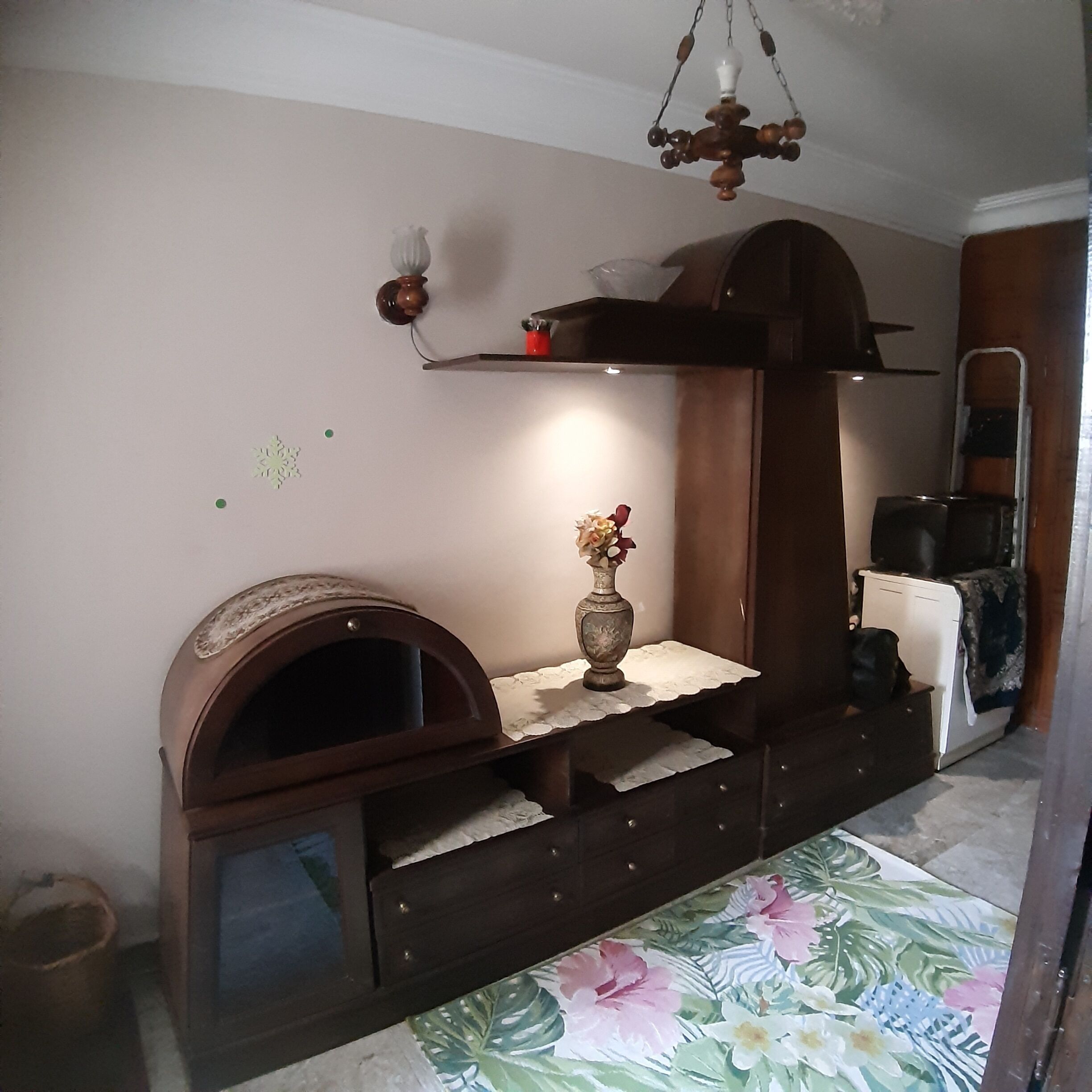 Een compacte kamer met een inbouwoven, een klein plankje met een vaas, een spiegel en een vloerkleed met bloemenprint.