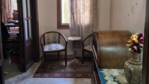 Una habitación con piso de madera, un tapete estampado, una silla de madera, una mesita redonda pequeña, un sofá, un jarrón decorativo y un ventilador de techo.