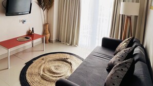 A modern living room with a sofa, a flat-screen TV mounted on the wall, a small table with a remote control, a floor lamp, and a large window with curtains. - Casa Kuta View 2 BR (Kuta)